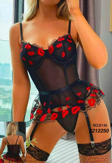 A woman modeling a sheer black lingerie bodysuit with red lip print and red floral applique, paired with black lace garter stockings in an indoor setting.